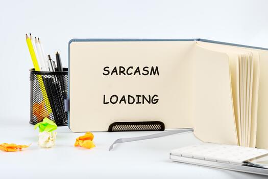 An open notebook displays the words Sarcasm Loading in bold letters. A pen holder filled with writing tools and crumpled paper sit nearby, creating a creative workspace setting photo