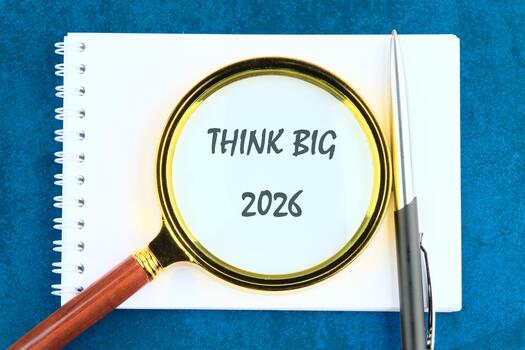 A notebook displays Think Big and 2026, emphasized by a magnifying glass, along with a pen resting on top, set against a textured, dark-blue background photo