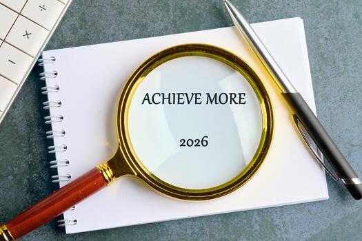 A notepad sits on a desk. On it sits a pen and a magnifying glass which is focused on the words Achieve More and the year 2026. A calculator is to the side photo