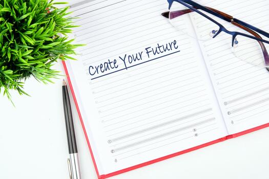 An open notebook displays Create Your Future written at the top of a page. Glasses rest on the right page, and a pen and small plant are next to the notebook on a clean, white surface photo