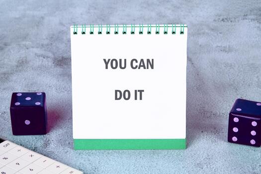 A notebook displaying the phrase YOU CAN DO IT sits on a grey surface next to a calculator and dice. An inspirational message is being portrayed in a still life photo