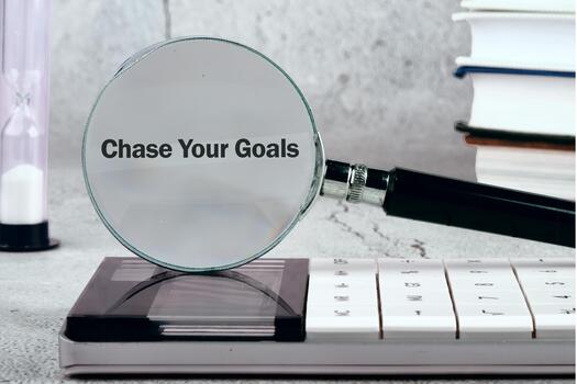 A magnifying glass highlights the phrase Chase Your Goals resting on a calculator, with an hourglass and a stack of books in the background. Focus on pursuing ambition photo