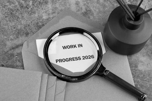 A magnifying glass rests on an envelope, focusing on a small white paper with the words Work in Progress 2026. An aroma diffuser sits nearby on a textured surface photo