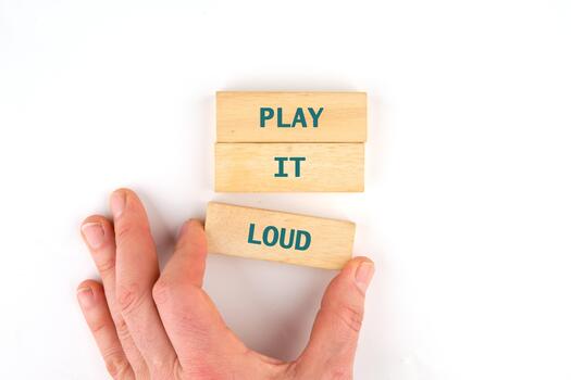 A hand places a wooden block labeled LOUD next to blocks spelling PLAY IT. The blocks are stacked against a white backdrop, suggesting a game or playful message photo