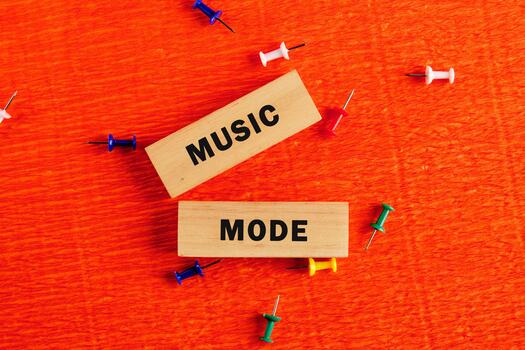 On a textured orange surface, two wooden blocks displaying the text MUSIC MODE are surrounded by colorful push pins, suggesting a creative and playful invitation to immerse oneself in music photo