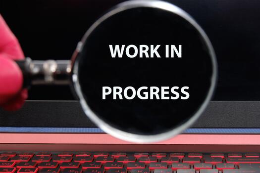 Magnifying glass focuses on the text WORK IN PROGRESS, viewed on a screen above a red illuminated keyboard. An unseen hand is holding the magnifying glass photo