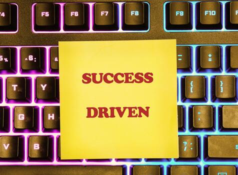 A yellow sticky note with the phrase SUCCESS DRIVEN lies on top of a multicolored, backlit keyboard, creating a visually striking message of motivation in a modern office photo