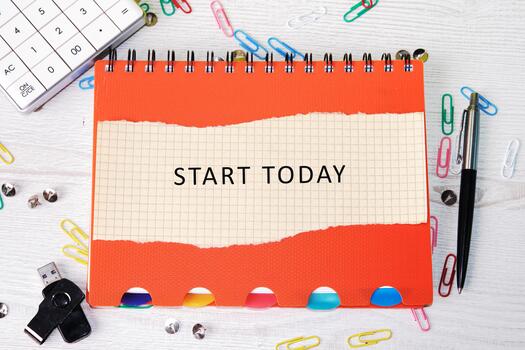 A spiral notebook, open to a page that reads START TODAY, sits on a desk with a pen, calculator, usb drive, push pins, and paper clips scattered around it photo