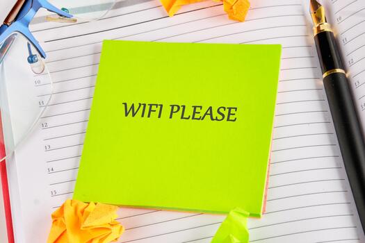 A green sticky note with the words WIFI PLEASE sits on a planner. A pen, glasses, and origami shapes surround it, conveying a message of urgency and a need for digital access photo