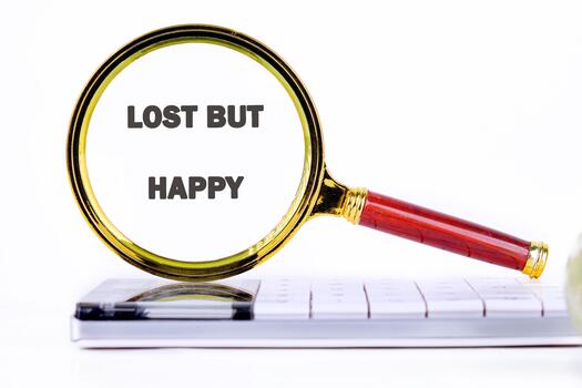 A magnifying glass with a red handle and gold rim shows the words lost but happy. It rests on top of a white computer keyboard. The background is plain white photo