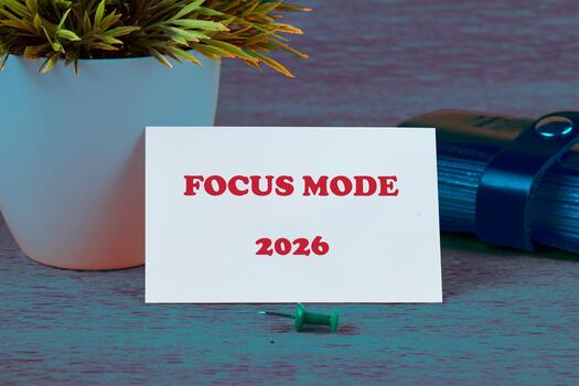 A notecard with FOCUS MODE and 2026 printed on it rests on a textured surface, propped up by a green pushpin, with a potted plant and a stack of books in the scene photo