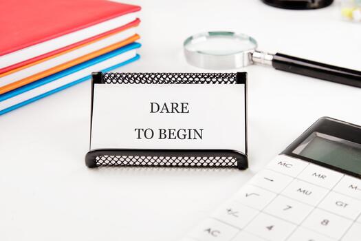 Card in a holder says DARE TO BEGIN near notebooks, a magnifying glass, and calculator on a white table. It inspires one to take a step forward and start something new photo