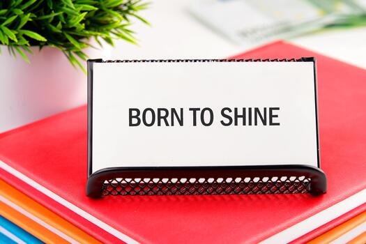 An inspiring message, Born To Shine, is displayed prominently on a desk. A stack of colorful books sit beneath it, next to a lively green plant, creating a bright and cheerful scene photo