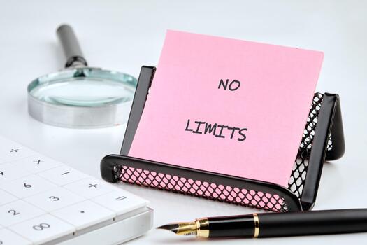 A pink note card reading NO LIMITS stands near a calculator, magnifying glass, and a fountain pen on a white surface, suggesting infinite possibilities and calculations photo