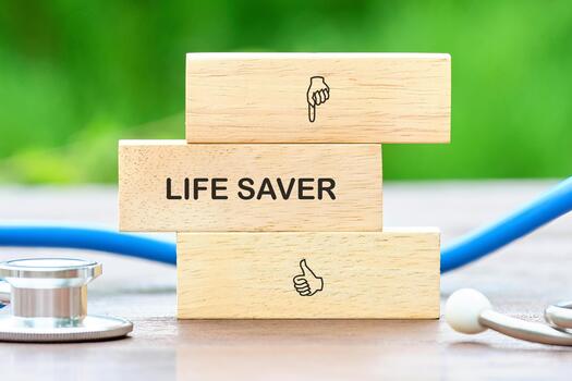 Three wooden blocks are stacked, with the middle block showing the words LIFE SAVER. The top and bottom blocks feature pointing and thumbs up icons. A stethoscope is nearby photo