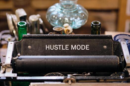 An old Typewriter displays the words HUSTLE MODE. It sits among old bottles, glass containers, and various metal objects on a wooden surface. A vintage store setting is suggested photo