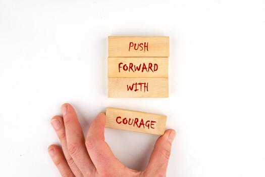 A hand reaches for wooden blocks arranged to read PUSH FORWARD WITH COURAGE. The blocks symbolize motivation and determination in a bright, inspiring workspace photo