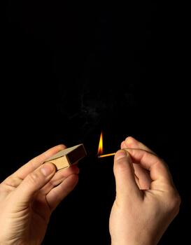 Hands holds burning match on a black background. A wooden match burns in the hands of a macro. Igniting a match on a box. Smoke from a match. Flash photo