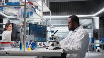 microbiologista dentro laboratório inspecionando espécime deslizar usando ótico dispositivo, analisando em pc. homem revendo citologia deslizar para clínico teste procedimento, introduzindo resultados em computador, Câmera uma video