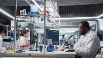 forskare rörelse förening mikroskop, håller på med prov observation i kontrollerade laboratorium miljö. afrikansk amerikan man studier vävnad under optisk enhet i bioteknik labb, kamera en video