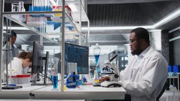 labb forskare använder sig av mikroskop till utvärdera cellulär strukturer. afrikansk amerikan man konstaterar prover genom förstoring verktyg på laboratorium bänk, framåt vetenskaplig kunskap, kamera en video