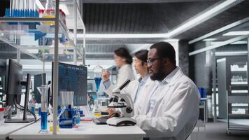 kemisk ingenjör använder sig av mikroskop till utvärdera testa prover i labb arbetsyta. forskning anläggning expert- konstaterar klinisk material genom förstoring verktyg på laboratorium bänk, kamera en video