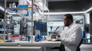 medicinsk laboratorium tekniker använder sig av vetenskaplig mikroskop för cellulär analys, håller på med forskning uppgifter. afrikansk amerikan man håller på med prov undersökning inuti labb med optisk enhet redskap, kamera en video