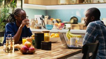 leende svart man arbetssätt avlägset på de tabell och chattar med flickvän, dricka kaffe medan de pojkvän skrivningar en Rapportera på hans dator. distansarbete på de frukost tabell. kamera b. video