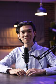Portrait of friendly talk show host seated at desk with microphone in cozy recording studio. Smiling asian man looking at camera while speaking into mic, podcasting and engaging online audience. photo