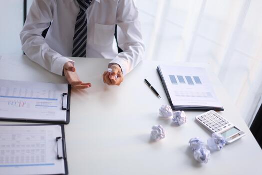 Man at office desk stressed or frustrated, crumpling paper and the presence of multiple crumpled paper balls on table. Concept for business weakness, creative block, or work pressure photo