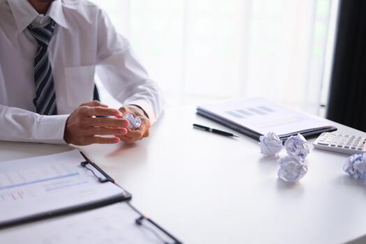 Man at office desk stressed or frustrated, crumpling paper and the presence of multiple crumpled paper balls on table. Concept for business weakness, creative block, or work pressure. photo