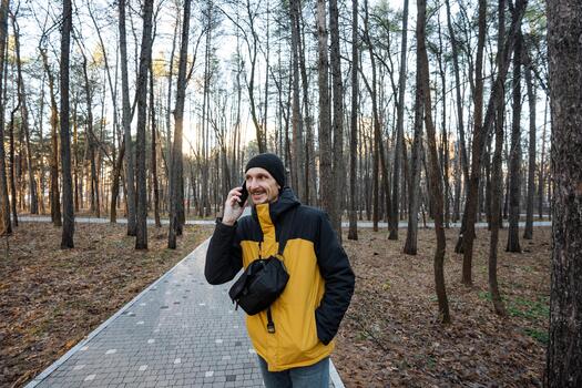 man walking through wooded path making phone call, crossbody bag and yellow jacket visible, steady stride, background of tall trees and paved trail, onthe-go communication mood photo