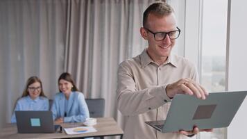 man bär glasögon innehar en bärbar dator stående nära fönster och har ringa upp. en par av kvinna kollegor sitta på skrivbord och skratt ser på dator på bakgrund i fläck. video