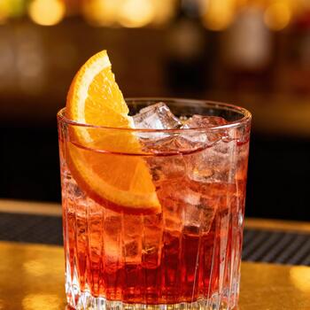 Chilled reddish beverage garnished with an orange slice sits in a textured glass on a bar surface photo