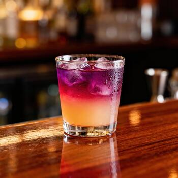 Vibrant layered cocktail with melting ice sits on a glossy wooden bar surface photo
