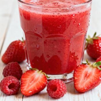 Bright red berry smoothie fills a glass surrounded by fresh whole and halved fruit on a light surface photo