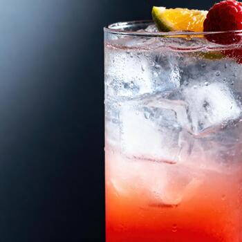 Tall glass filled with a chilled red beverage, ice cubes, and fruit garnish sits against a dark background. photo