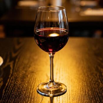 Stemmed glass filled with deep red beverage rests upon a richly illuminated wooden surface photo