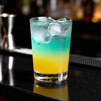 Brightly colored layered beverage rests on a polished dark bar surface with ice cubes photo