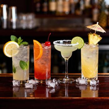 Four colorful, garnished mixed drinks are displayed on a dark wooden bar top. photo