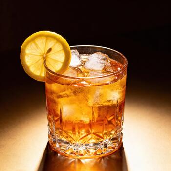 Amber colored beverage with ice and citrus garnish rests on illuminated surface photo