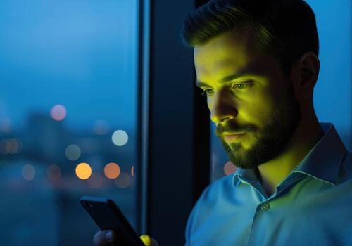 Focused professional man using smartphone by window at night with city lights photo