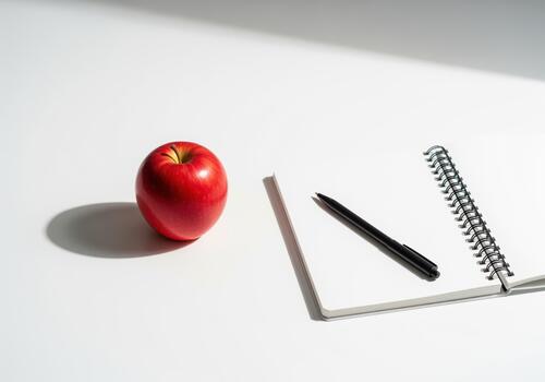 Vibrant red apple next to an open notebook and black pen on a clean white desk photo