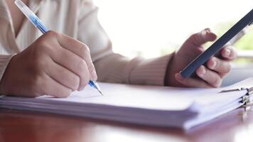 Hands writing on documents while holding a smartphone, showing multitasking, productivity, and everyday workflow with paperwork and digital technology, close-up video