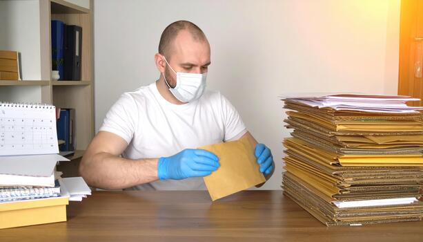 Man wearing protective face mask and blue gloves sorting brown envelopes at wooden desk with stacks of mail and calendar in office with warm sunlight photo