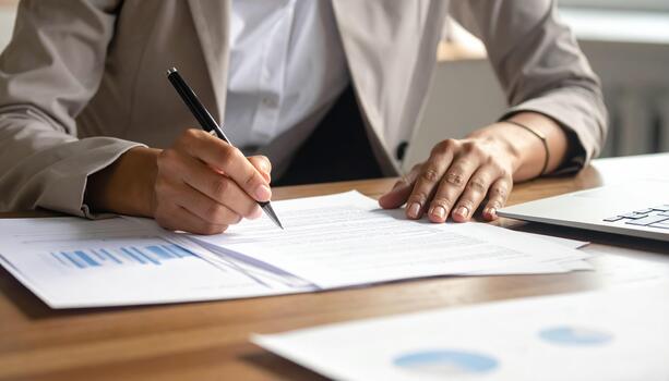 Person writing on document with pen at desk, business report and laptop nearby, focused on paperwork and analysis in office environment photo