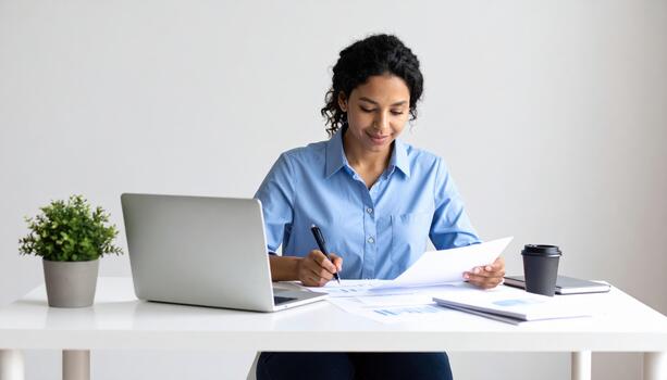 Woman is sitting at desk, focused on her work. She is reviewing documents and taking notes while using laptop. small plant and coffee cup are on table, creating productive atmosphere photo
