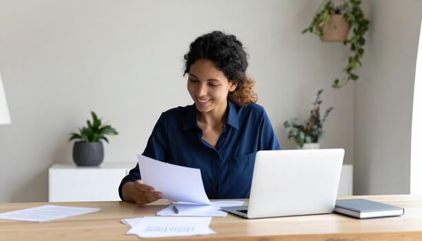 Woman is sitting wooden desk, smiling while reviewing documents. laptop is open front of her, and there are several papers scattered desk. Green plants are visible background, creating calm photo