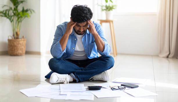 Man sits floor surrounded by papers, looking stressed and overwhelmed. He holds his head his hands, reflecting sense of anxiety and frustration. room is bright and minimalistic, with plant photo