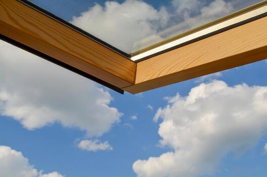 A wooden-framed skylight is open, showing a clear blue sky with fluffy white clouds through and around the window. photo